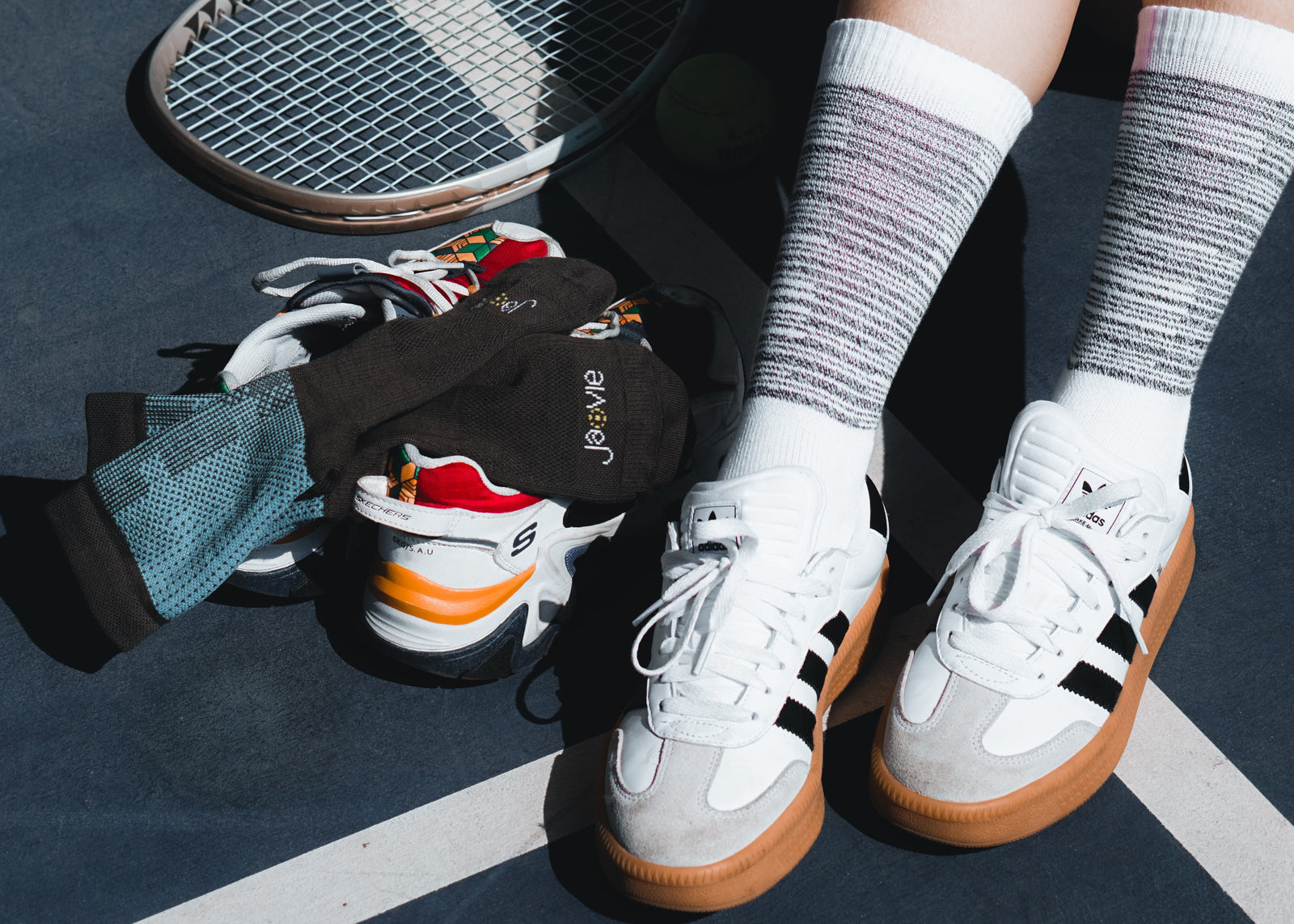 Person wearing white sneakers with brown soles on a tennis court, with a racket and shoes scattered around.